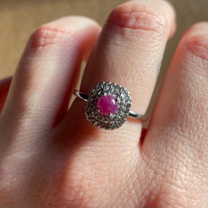 May include: A silver ring featuring a central, round, ruby gemstone surrounded by a square halo of smaller, clear stones. The ring's band is thin and polished, and the setting is detailed.