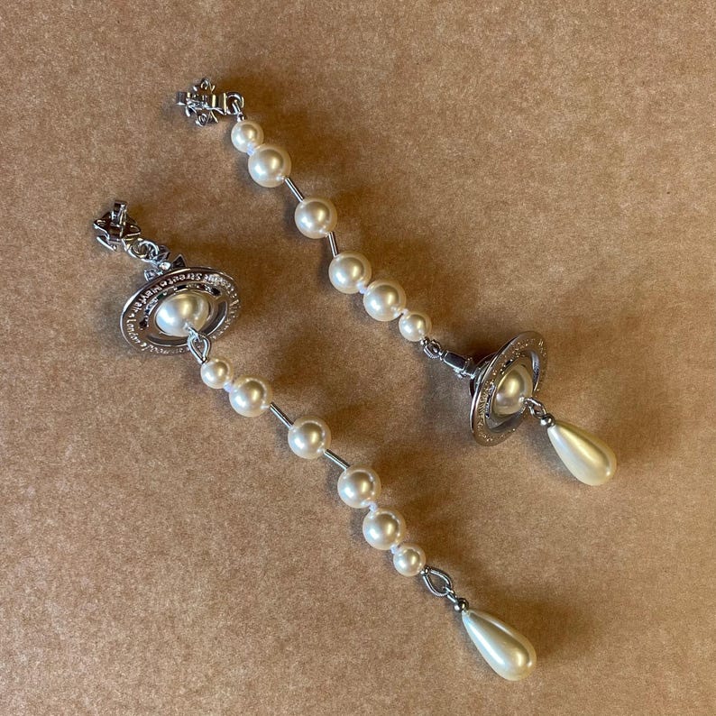 Pode incluir: Um par de brincos pendurados prateados com um design de p&eacute;rolas. Cada brinco tem um topo em forma de planeta com um centro de p&eacute;rola e uma p&eacute;rola pendurada em forma de gota. Os brincos s&atilde;o conectados por um fio de p&eacute;rolas.