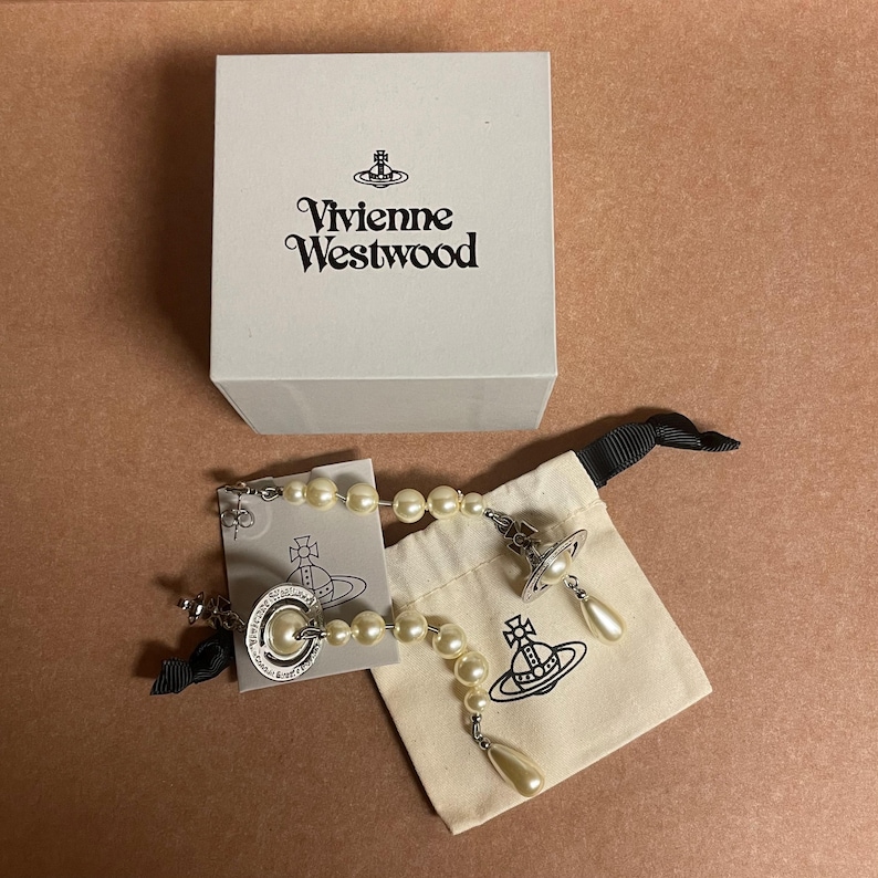 Pode incluir: Uma caixa branca com o logotipo da Vivienne Westwood est&aacute; posicionada acima de um par de brincos de p&eacute;rolas e uma pulseira a condizer. Os brincos e a pulseira t&ecirc;m um design de esfera prateada. Uma pequena bolsa branca suja com o logotipo da Vivienne Westwood tamb&eacute;m est&aacute; presente.