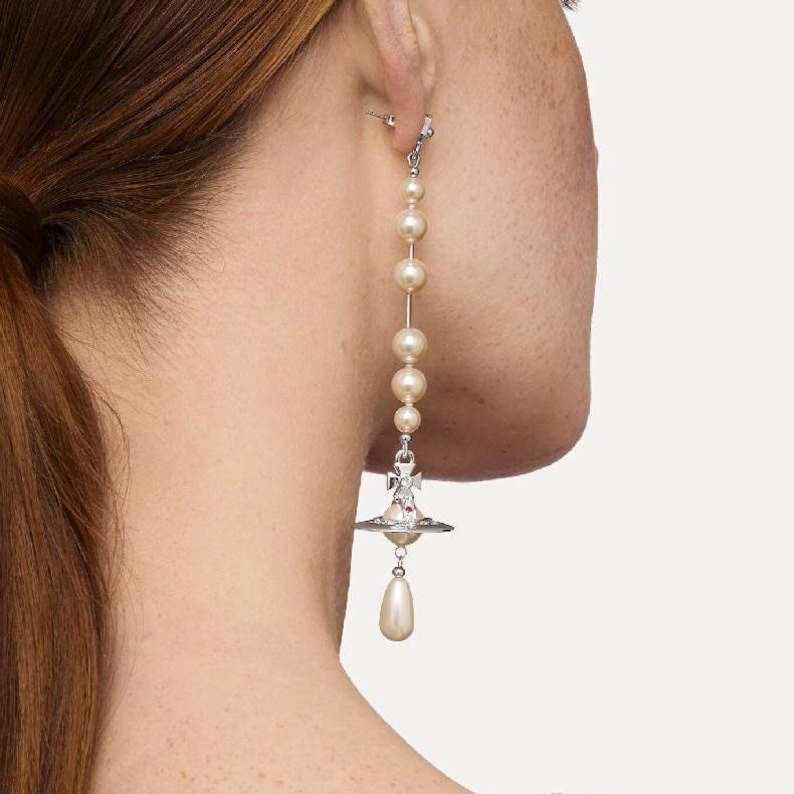 Può includere: Un orecchino lungo e pendente con perle color crema collegate da una catena d'argento. L'orecchino ha un ciondolo d'argento con una goccia di perla. L'orecchino è indossato sull'orecchio di una persona.