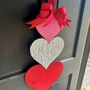 May include: Three wooden hearts in a stack, decorated for Valentine's Day. The top heart is pink with a red and white polka dot bow. The middle heart is white with a raised embossed pattern. The bottom heart is red.