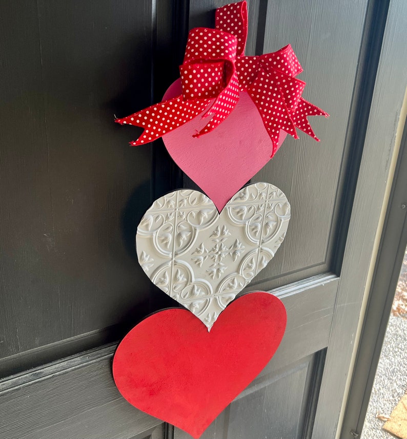 May include: Three wooden hearts in a stack, decorated for Valentine's Day. The top heart is pink with a red and white polka dot bow. The middle heart is white with a raised embossed pattern. The bottom heart is red.