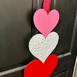 May include: A Valentine's Day door decoration with three hearts. The top heart is pink, the middle heart is silver with a textured pattern, and the bottom heart is red.