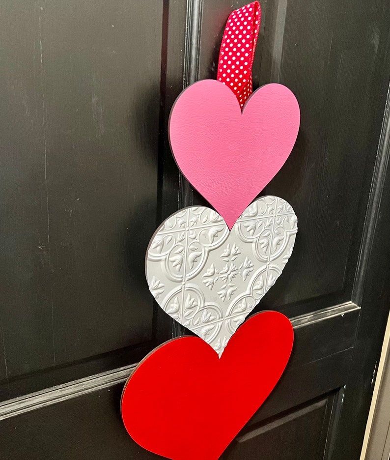 May include: A Valentine's Day door decoration with three hearts. The top heart is pink, the middle heart is silver with a textured pattern, and the bottom heart is red.
