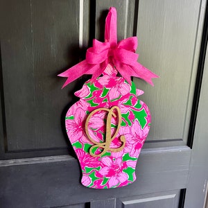 May include: A pink and green floral patterned wooden door hanger shaped like a Chinese ginger jar with a gold letter L on the front. A pink bow is attached to the top of the hanger.