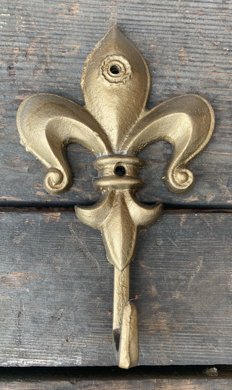 May include: Gold-colored metal fleur-de-lis hook. The hook is shaped like a stylized lily flower with a single hook at the bottom.