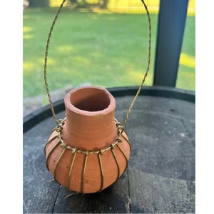 May include: A small terracotta pot with a gold wire handle and a gold wire cage around the base. The pot is sitting on a wooden surface.