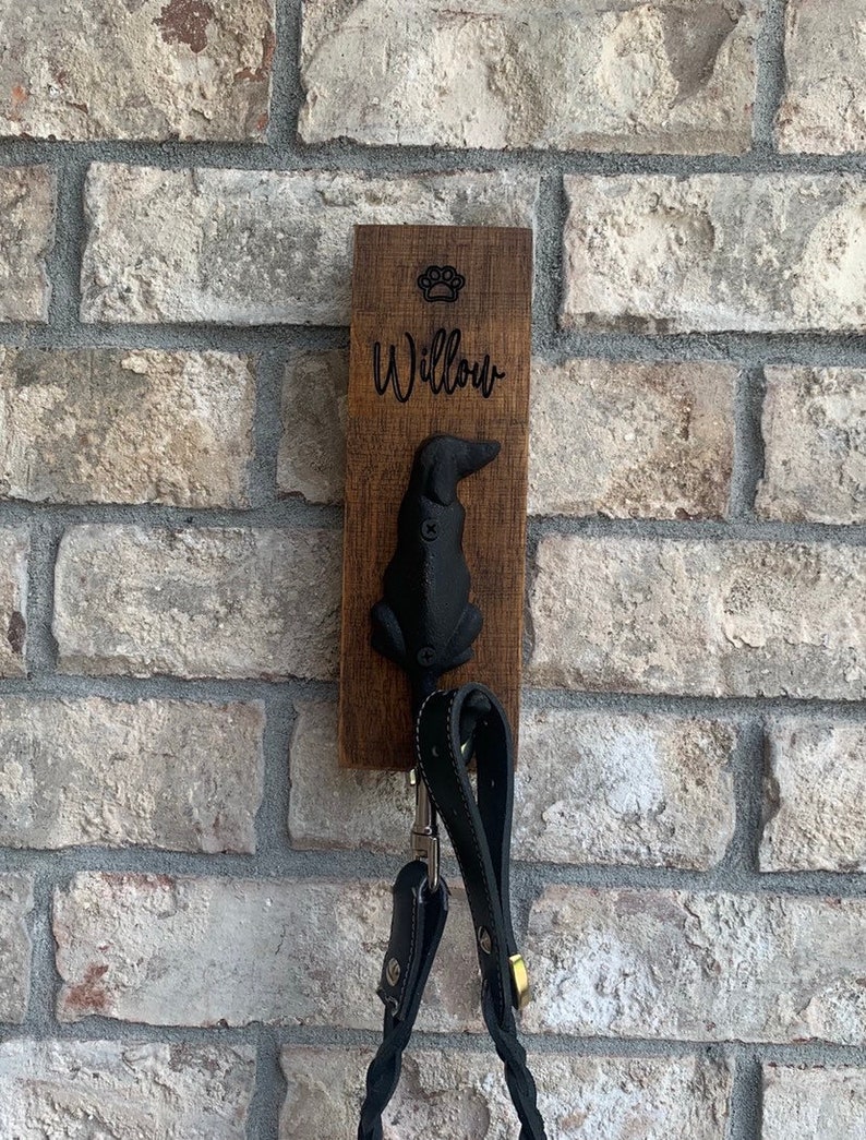 Op de afbeelding: Een houten hondenriemhaak met een zwart honden silhouet en de naam "Willow" erop gegraveerd. De haak is gemonteerd op een bakstenen muur en heeft twee riemen die eraan hangen.