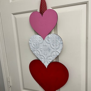 May include: Three wooden hearts in a vertical stack, with a pink heart on top, a white heart with a decorative pattern in the middle, and a red heart on the bottom.