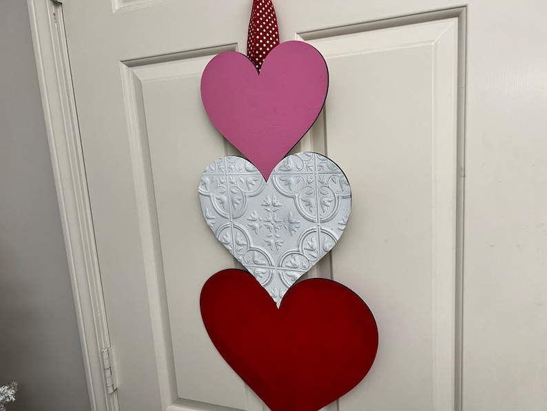 May include: Three wooden hearts in a vertical stack, with a pink heart on top, a white heart with a decorative pattern in the middle, and a red heart on the bottom.