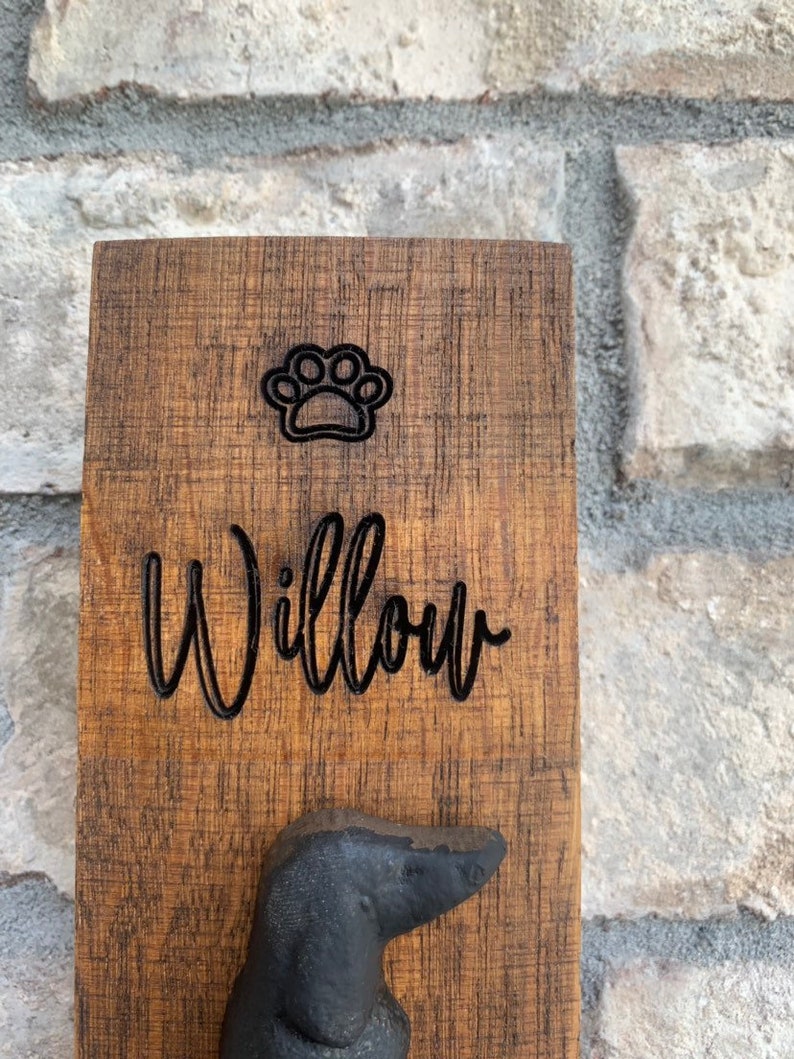 Op de afbeelding: Een houten hondenriemhaak met een pootprint en de naam "Willow" erop gegraveerd. De haak is in de vorm van een teckel.