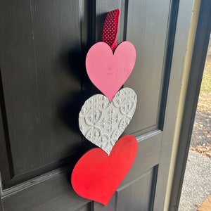 May include: A Valentine's Day door decoration with three hearts. The top heart is pink, the middle heart is white with a decorative pattern, and the bottom heart is red.