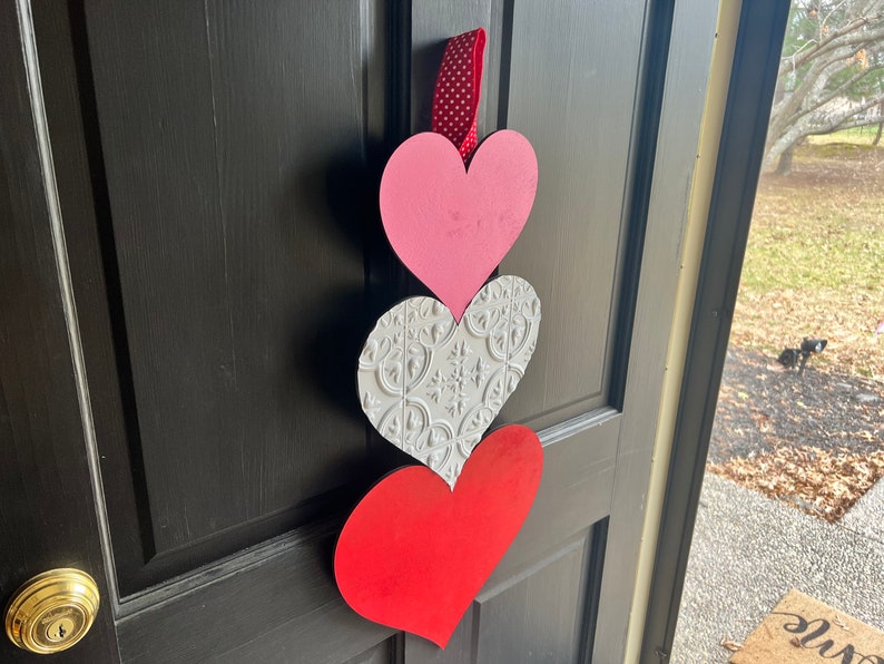 May include: A Valentine's Day door decoration with three hearts. The top heart is pink, the middle heart is white with a decorative pattern, and the bottom heart is red.