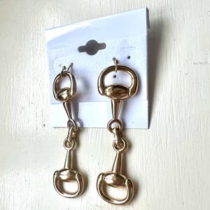 May include: A pair of gold-toned earrings with a horse bit design. The earrings are dangling and have a simple, elegant look.