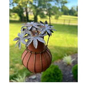 May include: A terracotta hanging planter with a wire cage and a cluster of metal flowers on top. The flowers are a silvery gray color with a rustic finish.