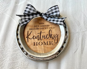 May include: A silver horseshoe ornament with a wooden insert. The insert has the text "The sun shines bright on my old Kentucky home" engraved on it. The horseshoe is decorated with a black and white checkered ribbon.