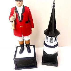 May include: A figurine of a man in a red jacket, black riding boots, and a black helmet holding a gold trumpet. Next to it is a black and white steeple-shaped figurine.