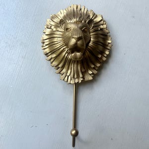 May include: A gold-colored lion head wall hook. The lion's mane is detailed with textured lines, and the hook extends downward with a small ball at the end. This decorative item is suitable for coats, hats, or other hanging items.
