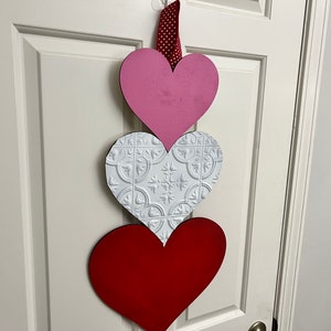 May include: Three wooden hearts hanging on a white door. The top heart is pink, the middle heart is white with a raised pattern, and the bottom heart is red.