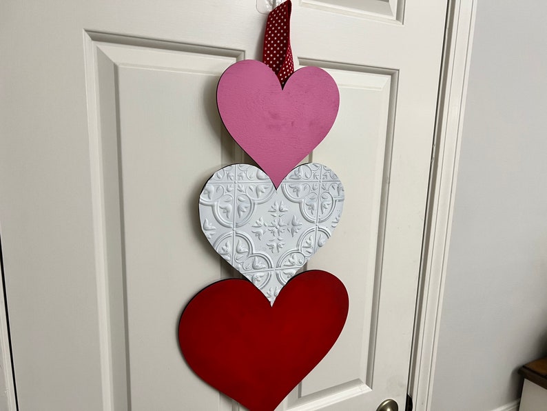 May include: Three wooden hearts hanging on a white door. The top heart is pink, the middle heart is white with a raised pattern, and the bottom heart is red.