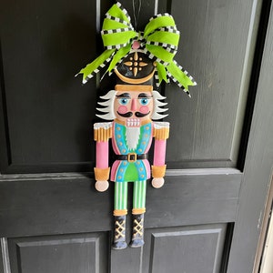 May include: A pink, green, and black nutcracker door hanger with a green and white bow. The nutcracker is wearing a pink jacket, green pants, and black boots.