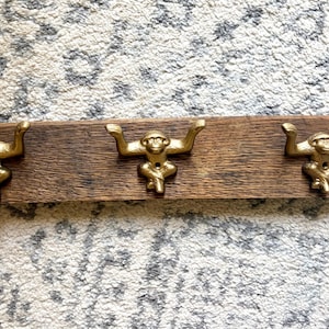 May include: A rectangular wooden wall hook rack featuring three gold-colored metal monkey figures. Each monkey is positioned with arms extended upward to serve as hooks. The wood has a natural brown grain and a curved top edge, providing a sturdy base for the three metallic monkey-shaped hooks.