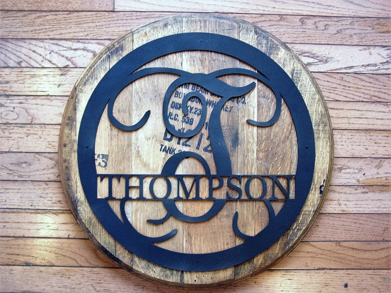Puede incluir: Un letrero redondo de madera con un monograma de metal negro de la letra "T" con el nombre "THOMPSON" debajo. El letrero est&aacute; hecho de un barril de whisky recuperado.