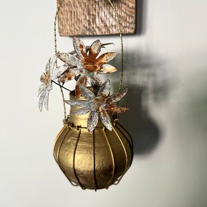 May include: A gold-painted metal vase with a wire cage design, hanging from a gold wire, with four rusty metal flowers.