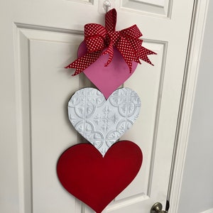 May include: A Valentine's Day door decoration with three hearts. The top heart is pink with a red and white polka dot bow. The middle heart is white with a raised embossed pattern. The bottom heart is red.
