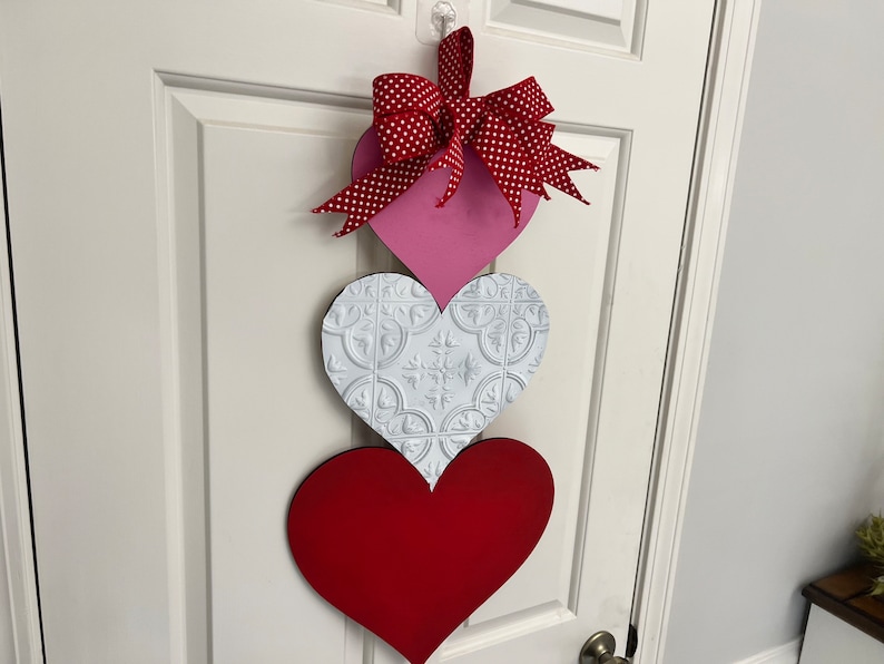 May include: A Valentine's Day door decoration with three hearts. The top heart is pink with a red and white polka dot bow. The middle heart is white with a raised embossed pattern. The bottom heart is red.