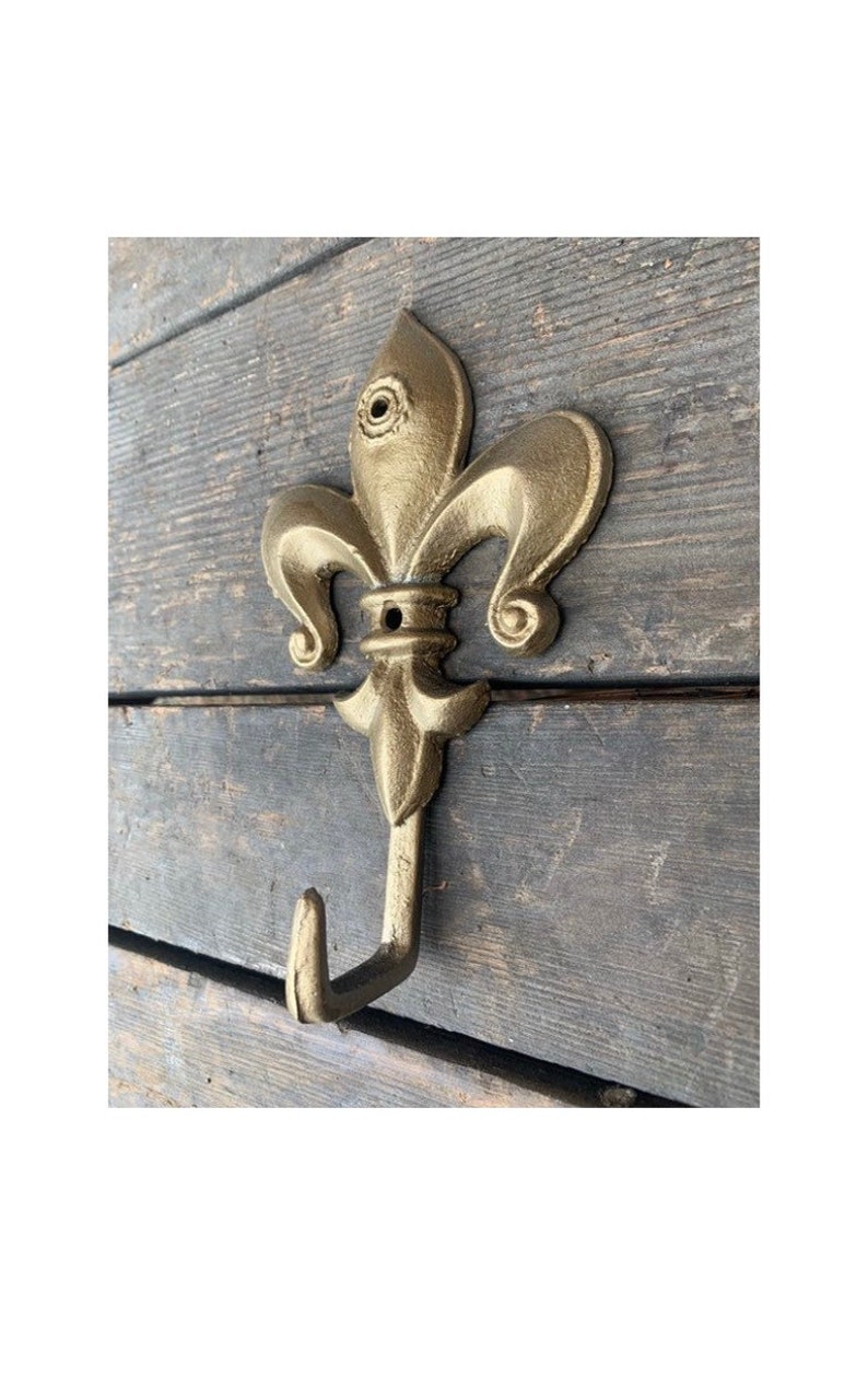 May include: A gold-colored metal hook with a fleur-de-lis design. The hook is mounted on a wooden surface.