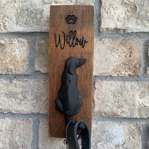 Op de afbeelding: Een houten hondenriemhaak met een zwart metalen hondensilhouet. De haak is gegraveerd met het woord "Willow" en een pootprint.