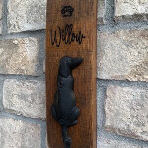 Op de afbeelding: Een houten bord met de naam "Willow" en een gegraveerde voetafdruk. Een zwarte metalen hondenhaak is bevestigd aan de onderkant van het bord.