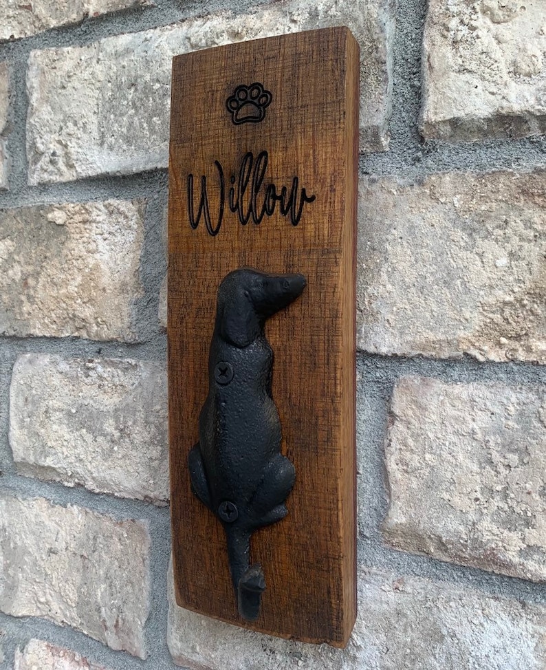 Op de afbeelding: Een houten bord met de naam "Willow" en een gegraveerde voetafdruk. Een zwarte metalen hondenhaak is bevestigd aan de onderkant van het bord.