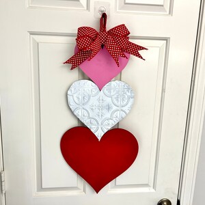 May include: A Valentine's Day door hanger with three hearts. The top heart is pink with a red and white polka dot bow. The middle heart is white with a raised pattern. The bottom heart is red.