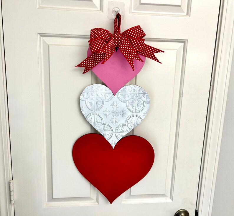 May include: A Valentine's Day door hanger with three hearts. The top heart is pink with a red and white polka dot bow. The middle heart is white with a raised pattern. The bottom heart is red.