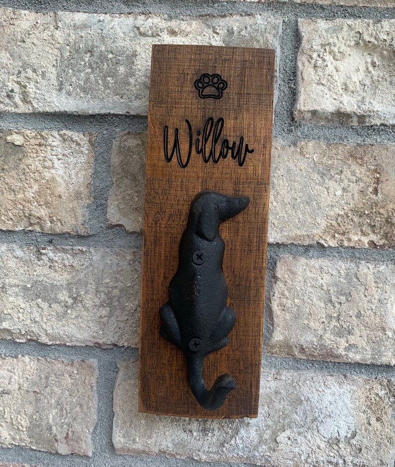 Op de afbeelding: Een houten hondenriemhaak met een zwart metalen hondensilhouet. De haak is gegraveerd met het woord "Willow" en een pootprint.