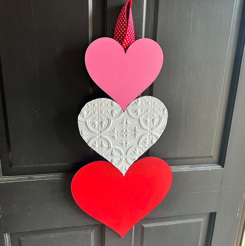 May include: A three-tiered heart-shaped door hanger. The top heart is pink, the middle heart is white with a decorative pattern, and the bottom heart is red.