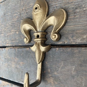 May include: A gold-colored metal hook with a fleur-de-lis design. The hook is mounted on a wooden surface.