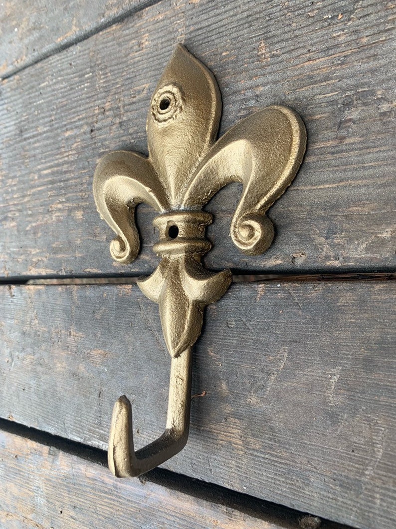 May include: A gold-colored metal hook with a fleur-de-lis design. The hook is mounted on a wooden surface.