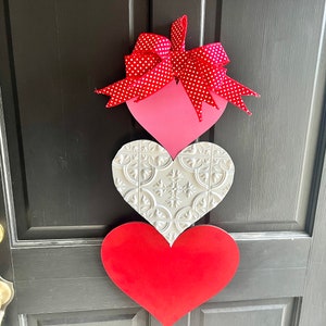 May include: A Valentine's Day door decoration with three wooden hearts. The top heart is pink with white polka dots and a red bow. The middle heart is white with a raised embossed pattern. The bottom heart is red.