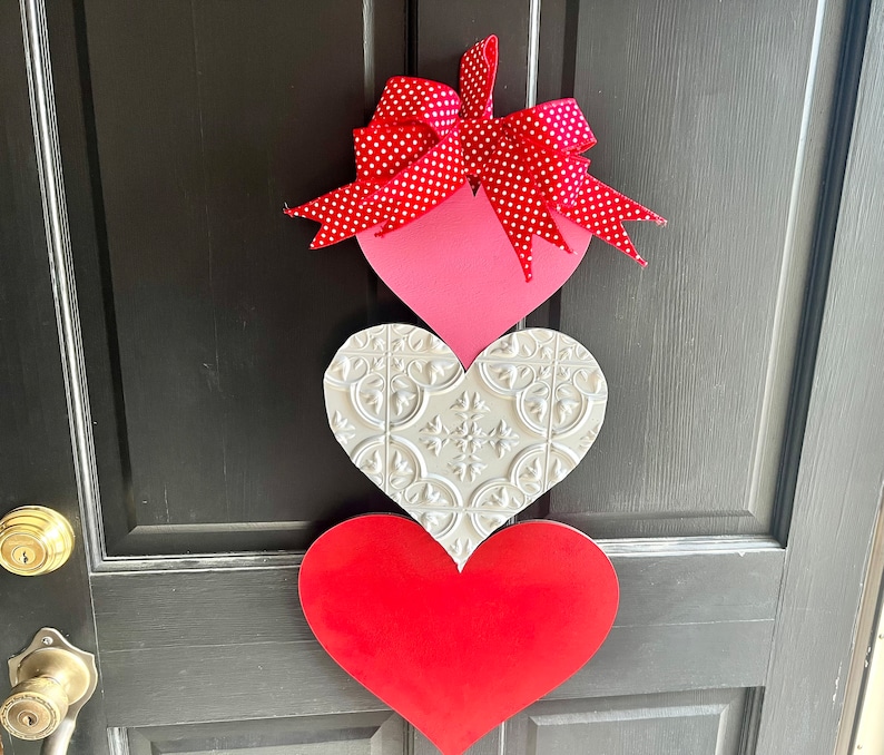 May include: A Valentine's Day door decoration with three wooden hearts. The top heart is pink with white polka dots and a red bow. The middle heart is white with a raised embossed pattern. The bottom heart is red.
