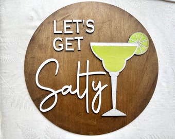 May include: A round wooden sign with the text "Let's Get Salty" in white lettering. A white margarita glass with a lime wedge is depicted on the sign.