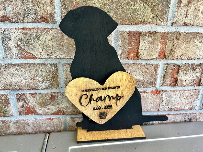 以下が含まれることがあります： 「Forever in our hearts Champ 2009 - 2022」と刻まれたハート型のプレートと、足跡がついた黒い木製の犬のシルエット