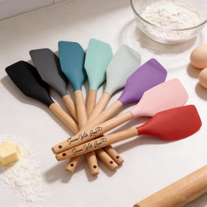 May include: A selection of silicone spatulas in various colours, each with a wooden handle. The colours include black, grey, teal, mint green, light grey, purple, pink, and red. The wooden handles are engraved with the text "Season With Love".