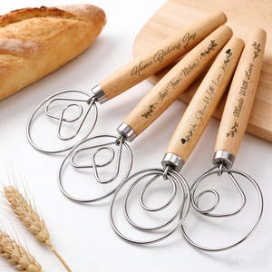 Puede incluir: Cuatro batidores de masa de acero inoxidable con mangos de madera. Cada mango está grabado con una frase diferente: "Home Baking Joy", "Love From Kitchen", "Slice With Love" y "Made With Love". Una barra de pan y espigas de trigo en el fondo.
