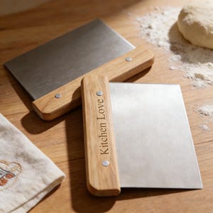 May include: Two stainless steel dough scrapers with wooden handles. One scraper is in front of the other, with the text "Kitchen Love" engraved on the handle. The scrapers are on a wooden surface, with a small cloth and flour in the background.