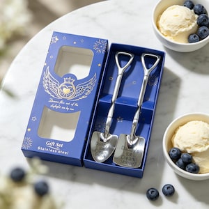 Könnte beinhalten: Ein Geschenkset aus zwei schaufelförmigen Edelstahllöffeln in einer blauen Schachtel mit goldenen Stern- und Flügelmotiven. Die Löffel sind mit dem Text Daddy's Ice Cream Shovel graviert. Daneben stehen zwei weiße Schalen mit Eis und Blaubeeren.