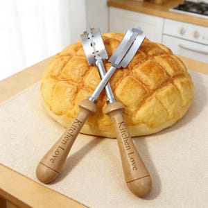 Op de afbeelding: Twee gekruiste broodmessen met houten handvatten, elk gegraveerd met "Kitchen Love". De zilveren messen liggen over een goudbruin, getextureerd brood. De gereedschappen zijn ontworpen om brood in te snijden.