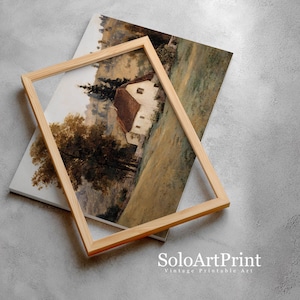 May include: A rectangular light-colored wooden frame rests on top of a landscape art print. The print depicts a white cottage with a brown roof nestled in a field with tall trees and rolling hills in muted earth tones of brown, green, and beige.